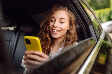 Genç bir kadın arabanın arka koltuğunda otururken akıllı telefon kullanır. Varış yerine giden yolda giderken arabada mutlu bir kadın. Teknoloji, blog yazma, iş konsepti.