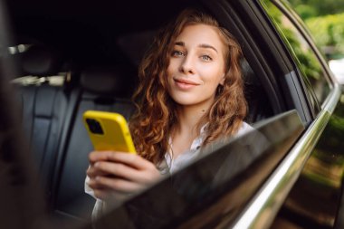 Genç bir kadın arabanın arka koltuğunda otururken akıllı telefon kullanır. Varış yerine giden yolda giderken arabada mutlu bir kadın. Teknoloji, blog yazma, iş konsepti.