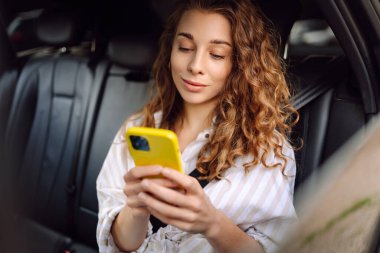Genç bir kadın arabanın arka koltuğunda otururken akıllı telefon kullanır. Varış yerine giden yolda giderken arabada mutlu bir kadın. Teknoloji, blog yazma, iş konsepti.