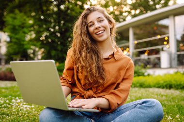 Yeşil çimlerin üzerinde dizüstü bilgisayarı olan genç kıvırcık kadın. Eğitim çevrimiçi. Bilgi işlem. İş, blog yazma, serbest çalışma kavramı.