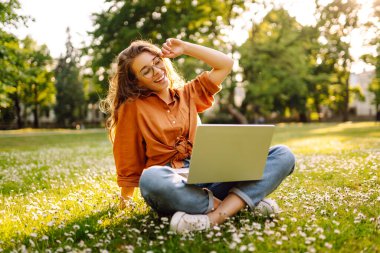 Yeşil çimlerin üzerinde dizüstü bilgisayarı olan genç kıvırcık kadın. Eğitim çevrimiçi. Bilgi işlem. İş, blog yazma, serbest çalışma kavramı.