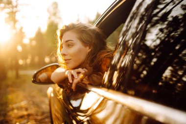 Genç bir kadın gün batımında bir arabanın camından sarkıyor. Yaşam tarzı, seyahat, turizm, doğa, aktif yaşam.
