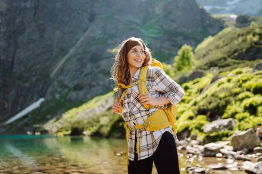 Sarı bir sırt çantasıyla dağlarda orman ve kayalıkların arasında yürüyüş parkurları boyunca seyahat eden genç bir kadın. Yaşam tarzı, macera, doğa, aktif yaşam.