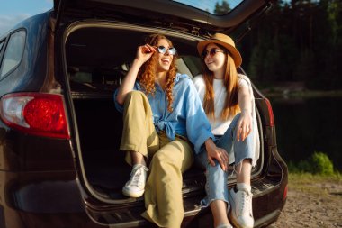 Seyahat ederken selfie. Mutlu kadınlar bir arabanın bagajında oturur ve akıllı telefon kullanır. Arabayla seyahat etme kavramı, aktif yaşam tarzı, blog yazma.
