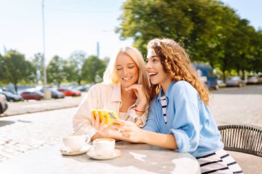 İki genç kadın kafede oturmuş, kahvelerini yudumluyor ve akıllı telefonlarıyla eğleniyorlar. Alışverişten sonra kahve molası. Moda, güzellik, blog, turizm.