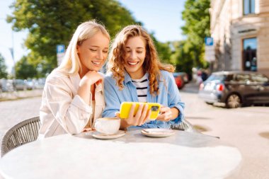 İki genç kadın kafede oturmuş, kahvelerini yudumluyor ve akıllı telefonlarıyla eğleniyorlar. Alışverişten sonra kahve molası. Moda, güzellik, blog, turizm.