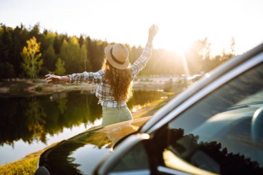 Güneşli bir günde arabanın yanında duran genç bir kadın, doğada arabayla seyahat ediyor, özgürlük. Etkin yaşam biçimi.