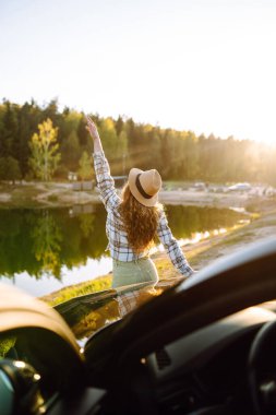 Güneşli bir günde arabanın yanında duran genç bir kadın, doğada arabayla seyahat ediyor, özgürlük. Etkin yaşam biçimi.