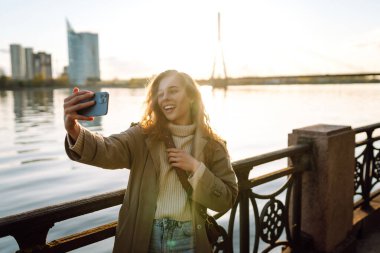 Selfie zamanı. Elinde cep telefonu olan genç bir kadın rıhtımda duran akıllı telefon kamerasıyla selfie çekiyor. Yaşam tarzı, seyahat, turizm, tatil, teknoloji, hafta sonu.