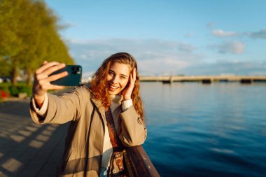 Selfie zamanı. Elinde cep telefonu olan genç bir kadın rıhtımda duran akıllı telefon kamerasıyla selfie çekiyor. Yaşam tarzı, seyahat, turizm, tatil, teknoloji, hafta sonu.