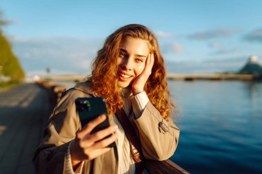 Selfie zamanı. Elinde cep telefonu olan genç bir kadın rıhtımda duran akıllı telefon kamerasıyla selfie çekiyor. Yaşam tarzı, seyahat, turizm, tatil, teknoloji, hafta sonu.