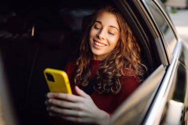 Bir arabanın arka koltuğunda elinde akıllı telefon olan kıvırcık bir kadının portresi. Teknoloji kavramı, araba ve iş seyahati..