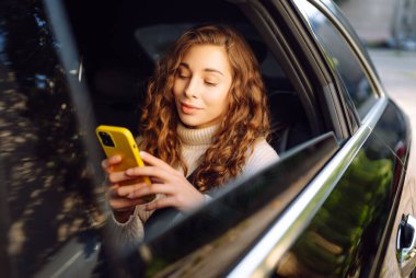 Bir arabanın arka koltuğunda elinde akıllı telefon olan kıvırcık bir kadının portresi. Teknoloji kavramı, araba ve iş seyahati..