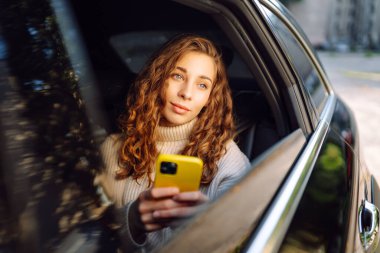 Bir arabanın arka koltuğunda elinde akıllı telefon olan kıvırcık bir kadının portresi. Teknoloji kavramı, araba ve iş seyahati..