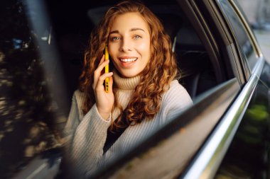 Bir arabanın arka koltuğunda elinde akıllı telefon olan kıvırcık bir kadının portresi. Teknoloji kavramı, araba ve iş seyahati..