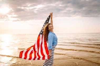 Gün batımında kumsalda Amerikan bayrağı taşıyan kadın vatansever. ABD 4 Temmuz 'u kutluyor. Bağımsızlık Günü kavramı