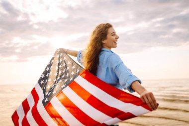 Gün batımında kumsalda Amerikan bayrağı taşıyan kadın vatansever. ABD 4 Temmuz 'u kutluyor. Bağımsızlık Günü kavramı