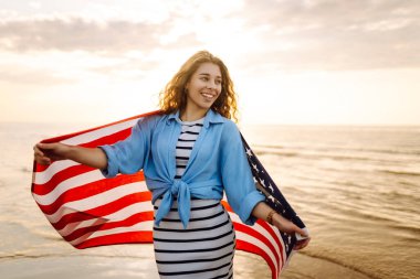 Gün batımında kumsalda Amerikan bayrağı taşıyan kadın vatansever. ABD 4 Temmuz 'u kutluyor. Bağımsızlık Günü kavramı