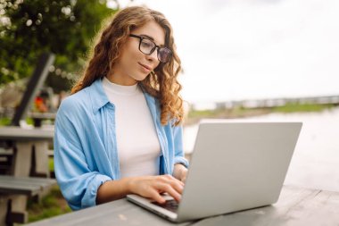 Parktaki bir masada dizüstü bilgisayar kullanarak oturan güzel bir kadın. Eğitim, iş, blog veya serbest çalışma kavramı.