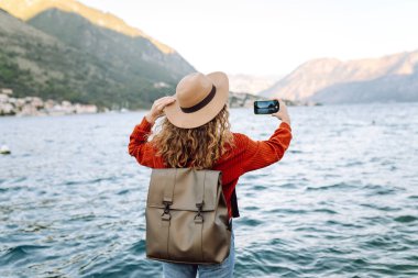 Sırt çantalı genç bir kadının rıhtımda beklerken dağ ve göl fotoğraflarını çekmek için akıllı telefon kullanışının arka planı. Yaşam tarzı, seyahat, turizm, aktif yaşam.