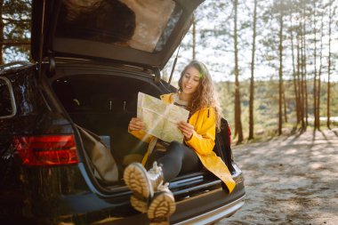 Mutlu kadın haritada yerini arıyor açık hava hatchback arabasının bagajında oturuyor. Boş zaman, yolculuk, seyahat ve insan konsepti.
