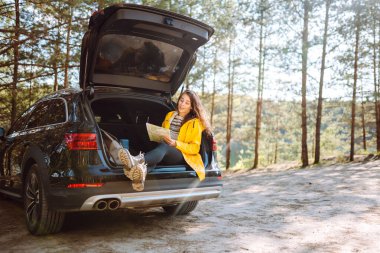 Mutlu kadın haritada yerini arıyor açık hava hatchback arabasının bagajında oturuyor. Boş zaman, yolculuk, seyahat ve insan konsepti.