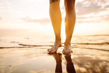 İnce kadın bacakları ve ayakları kumlu sahilde deniz suyu dalgaları boyunca yürüyor. Güzel bir kadın deniz kıyısında sörf yapıyor. Rahatlama, seyahat, özgürlük ve yaz tatili kavramı..