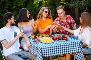 Bir grup arkadaş bira fabrikasının bahçesinde bira bardaklarını tokuşturuyor. Tatil, piknik, hafta sonu, doğa.