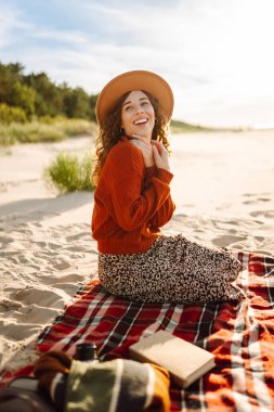 Samimi bir kazak ve şapka giyen genç bir kadın sahilde bir battaniyede otururken kitap okuyarak doğanın tadını çıkarıyor. Tatil konsepti, hafta sonu. Yaşam biçimi.