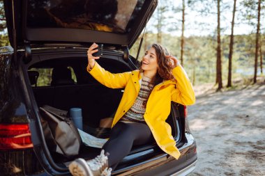 Kadın arabanın bagajında dinleniyor ve selfie çekiyor. Yaşam tarzı, seyahat, turizm, doğa, aktif yaşam.