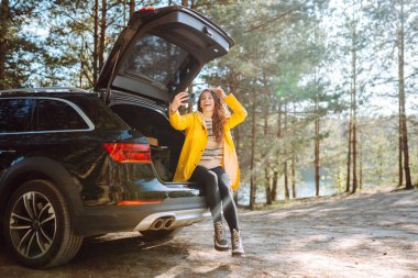 Kadın arabanın bagajında dinleniyor ve selfie çekiyor. Yaşam tarzı, seyahat, turizm, doğa, aktif yaşam.