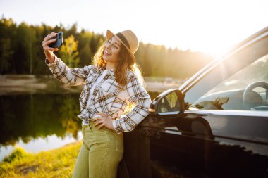 Arabayla seyahat eden genç bayan turist arabanın yanında selfie çekiyor. Aktif yaşam tarzı, blog yazma, doğa, hafta sonu.