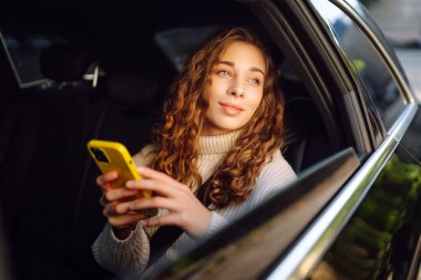 Genç bir kadın arabanın arka koltuğunda oturur ve telefonda konuşur. İş, teknoloji, seyahat, online iletişim kavramı. Taksi..
