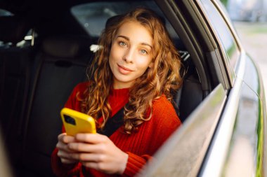 Genç bir kadın arabanın arka koltuğunda oturur ve telefonda konuşur. İş, teknoloji, seyahat, online iletişim kavramı. Taksi..