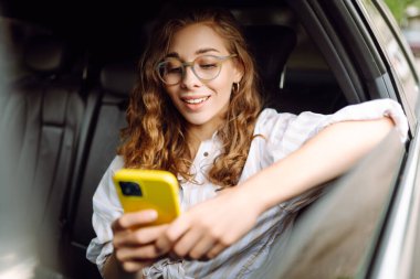Bir arabanın arka koltuğunda elinde akıllı telefonuyla mutlu bir kadın. Teknoloji kavramı, araba ve iş seyahati..