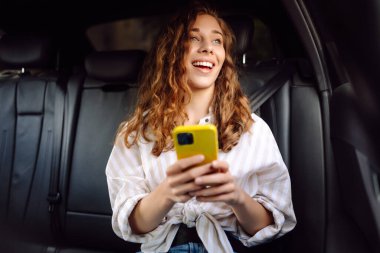 Bir arabanın arka koltuğunda elinde akıllı telefonuyla mutlu bir kadın. Teknoloji kavramı, araba ve iş seyahati..
