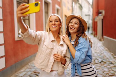 İki güzel genç bayan arkadaş şehirde birlikte yürüyüp, selfie çekerken akıllı telefon kullanıyorlar. Moda, seyahat, teknoloji, blog konsepti.
