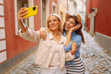 İki güzel genç bayan arkadaş şehirde birlikte yürüyüp, selfie çekerken akıllı telefon kullanıyorlar. Moda, seyahat, teknoloji, blog konsepti.