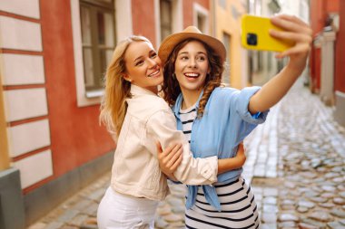 İki güzel genç bayan arkadaş şehirde birlikte yürüyüp, selfie çekerken akıllı telefon kullanıyorlar. Moda, seyahat, teknoloji, blog konsepti.