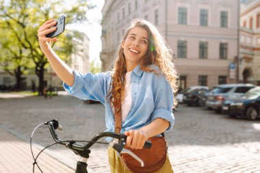 Şehir sokaklarında bisiklet sürerken cep telefonu kullanan genç bir kadın. Telekomünikasyon, video konferans, aile ve arkadaşlarla uzaktan iletişim. Selfie zamanı..