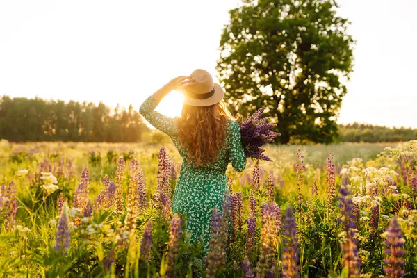 Elinde bir demet lavanta çiçeği tutan güzel bir kadın yaz çayırlarında yürüyor. Moda, güzellik, doğa konsepti. Şifalı bitkilerin toplanması.