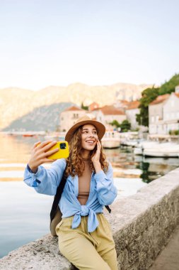 Genç bir kadın sokakta yürüyor ve akıllı telefon kamerasıyla selfie çekiyor. Olumlu anlar kavramı, eğlence. Turizm ve teknoloji