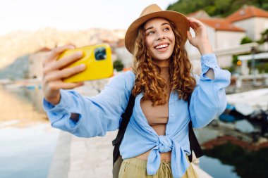 Genç bir kadın sokakta yürüyor ve akıllı telefon kamerasıyla selfie çekiyor. Olumlu anlar kavramı, eğlence. Turizm ve teknoloji