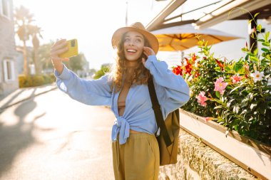 Genç bir kadın sokakta yürüyor ve akıllı telefon kamerasıyla selfie çekiyor. Olumlu anlar kavramı, eğlence. Turizm ve teknoloji