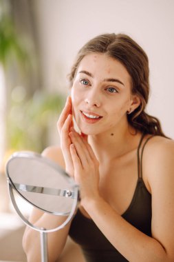 Güzel bir kadın aynaya bakıyor, eliyle yüzüne dokunuyor, sivilceleri ve pigmentasyonu inceliyor. Tıp ve kozmetoloji.