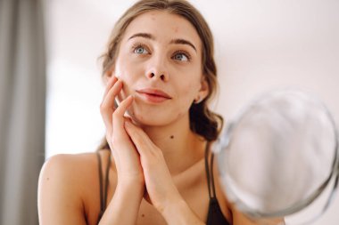 Güzel bir kadın aynaya bakıyor, eliyle yüzüne dokunuyor, sivilceleri ve pigmentasyonu inceliyor. Tıp ve kozmetoloji.