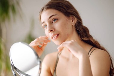 Güzel bir kadın aynaya bakıyor, eliyle yüzüne dokunuyor, sivilceleri ve pigmentasyonu inceliyor. Tıp ve kozmetoloji.