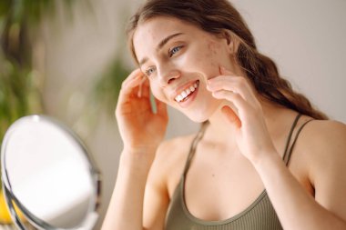 Güzel bir kadın aynaya bakıyor, eliyle yüzüne dokunuyor, sivilceleri ve pigmentasyonu inceliyor. Tıp ve kozmetoloji.