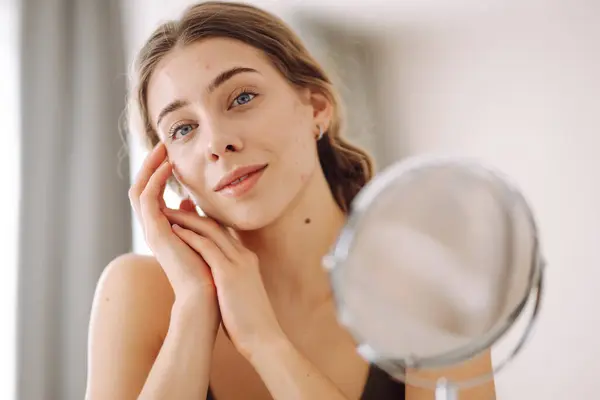 stock image Beautiful woman looking in the mirror, touching her face with her hand, examines acne, pigmentation. Medicine and cosmetology.