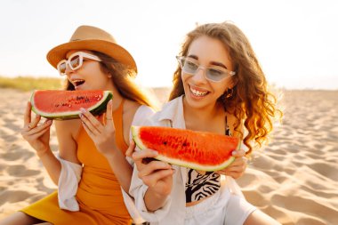 Yaz tatilinde, arka planda deniz olan bir karpuz yiyen, gün batımında kumsalda oturan dişi arkadaşlar. İnsanlar, yaşam tarzı, seyahat, doğa ve tatil kavramı.